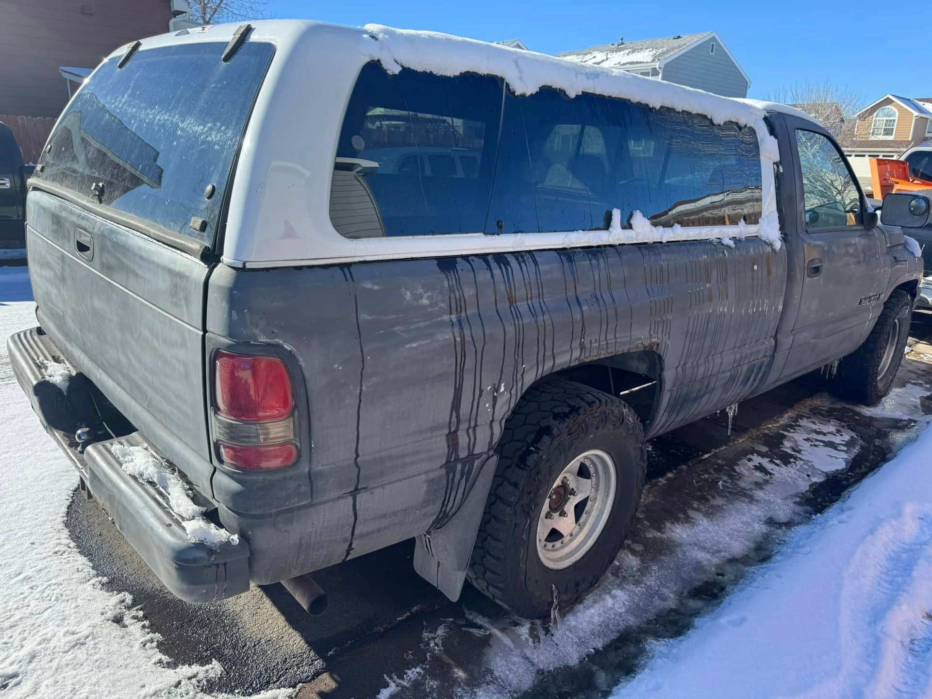 Dark pickup truck with white camper shell covered in snow, parked in snowy driveway.