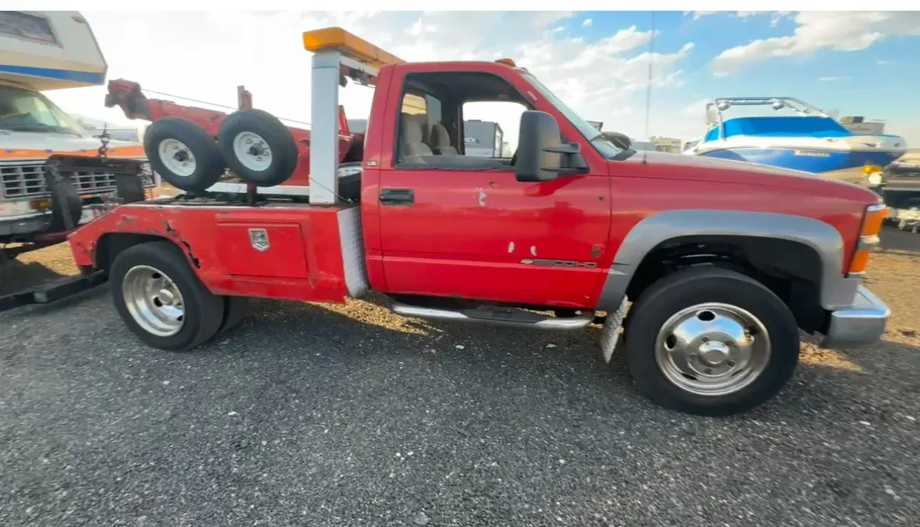 Red tow truck parked on gravel, with a tow arm and spare tires visible.