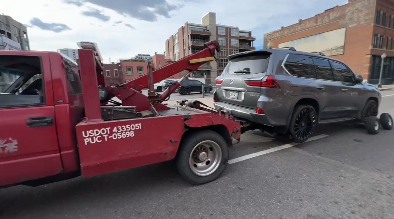 A red tow truck towing a gray SUV on a city street; buildings in the background.