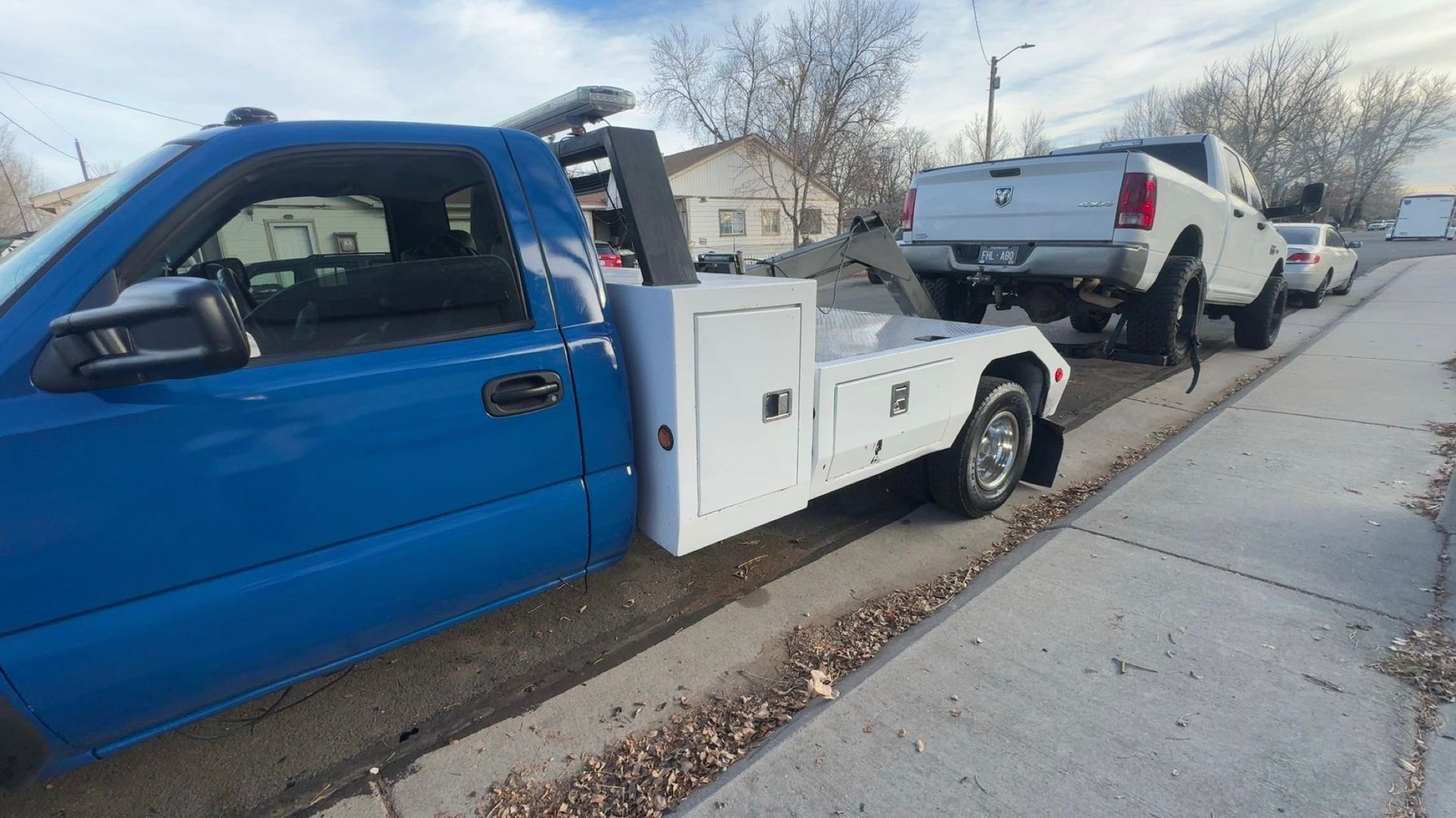 Blue tow truck towing a white pickup truck on a city street.