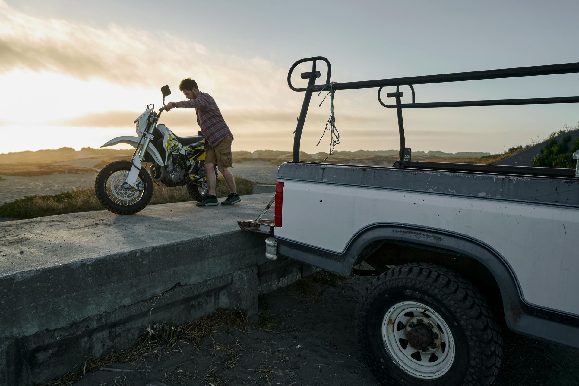 Man pushing a dirt bike near a truck parked by a coastal road at sunset.