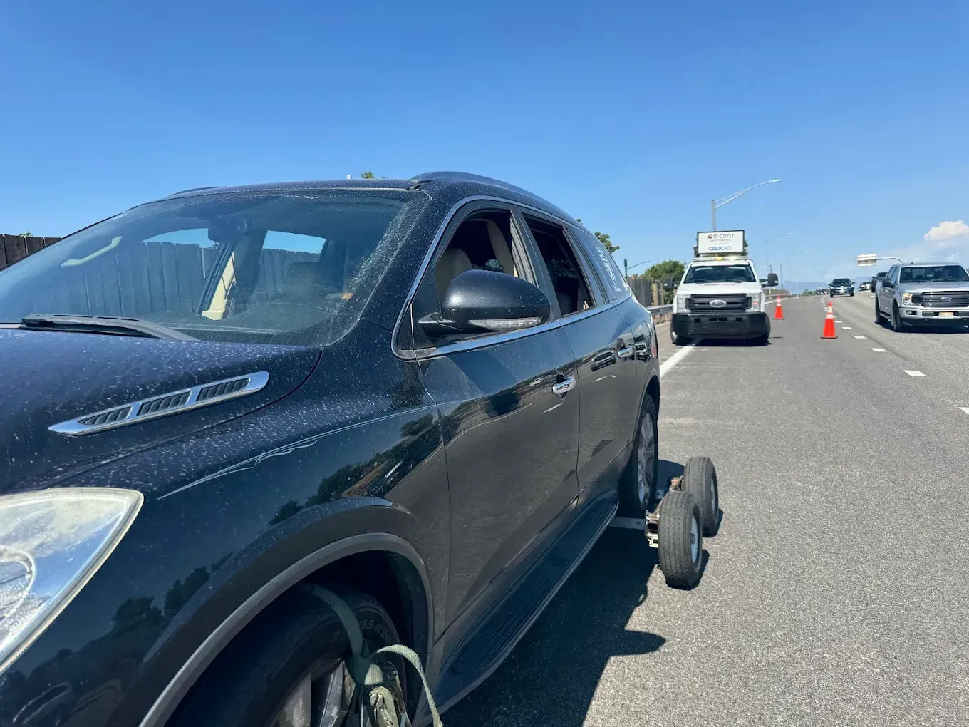 Black SUV being towed on a highway with two wheels removed. Utility vehicles and cones in background.