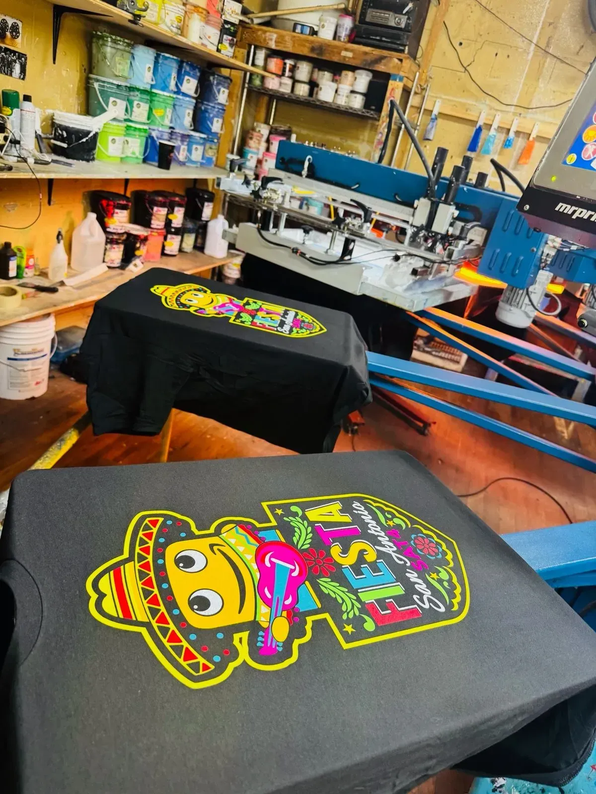 Two black t-shirts with colorful cartoon designs, near a screen printing machine in a workshop.