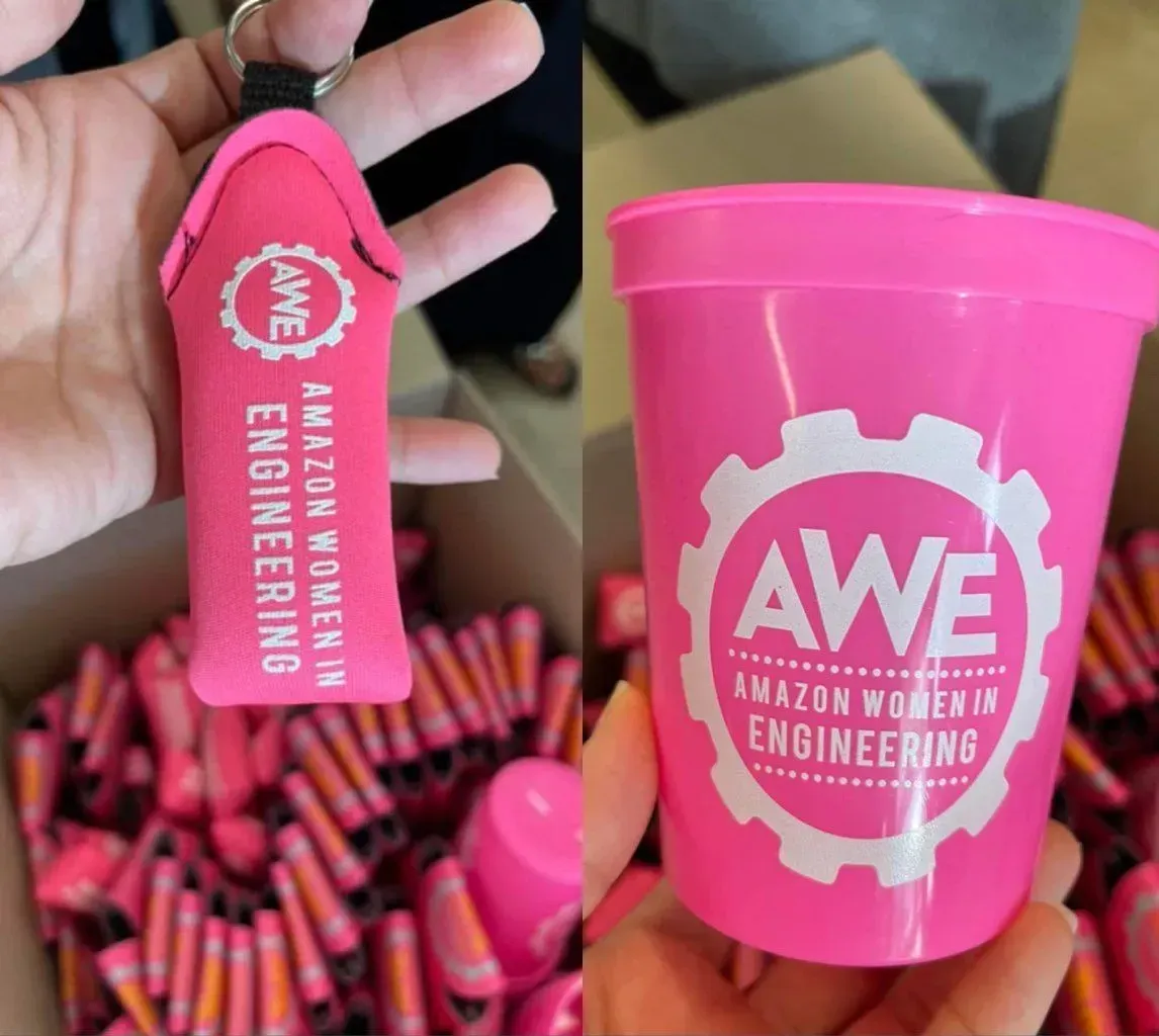 Two pink Amazon Women in Engineering giveaways: a keychain holder and a cup, with the AWE logo.