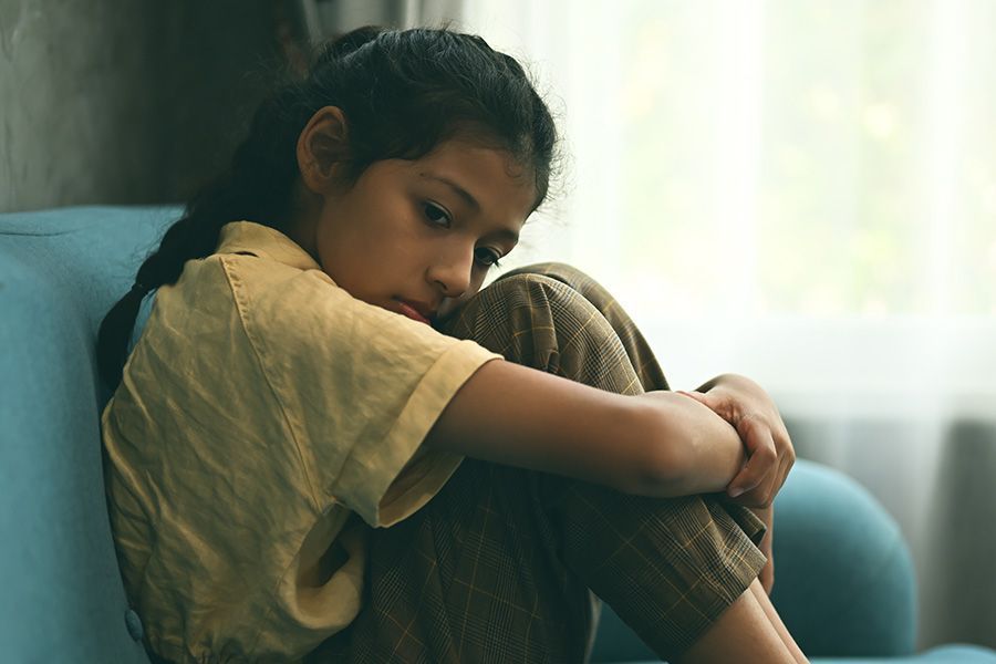 A young girl is sitting on a blue couch hugging her knees.