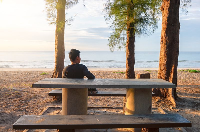 A man is sitting at a picnic table on the beach looking at the ocean.