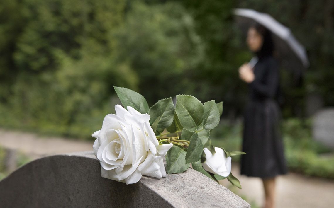 A white rose is sitting on a gravestone next to a woman holding an umbrella.