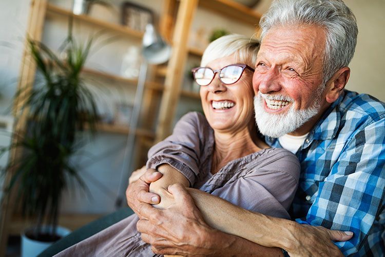 An elderly couple is hugging each other and smiling while sitting on a couch.