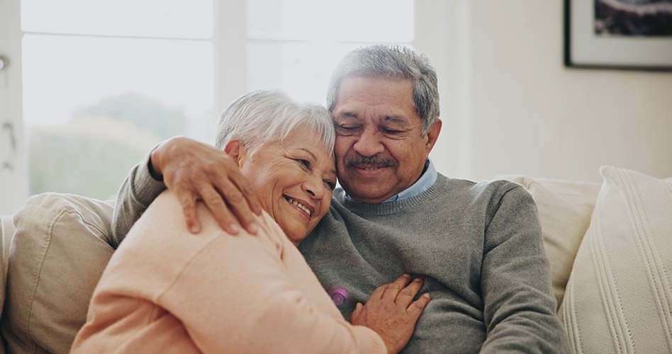 An elderly couple is sitting on a couch hugging each other.