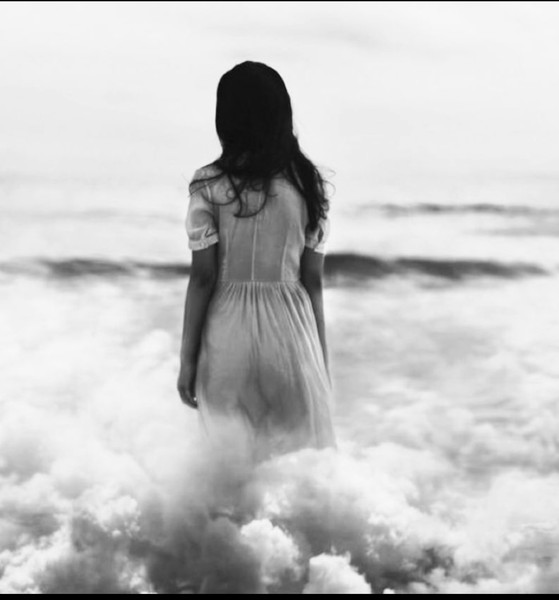 A woman in a white dress is standing in the ocean surrounded by clouds.