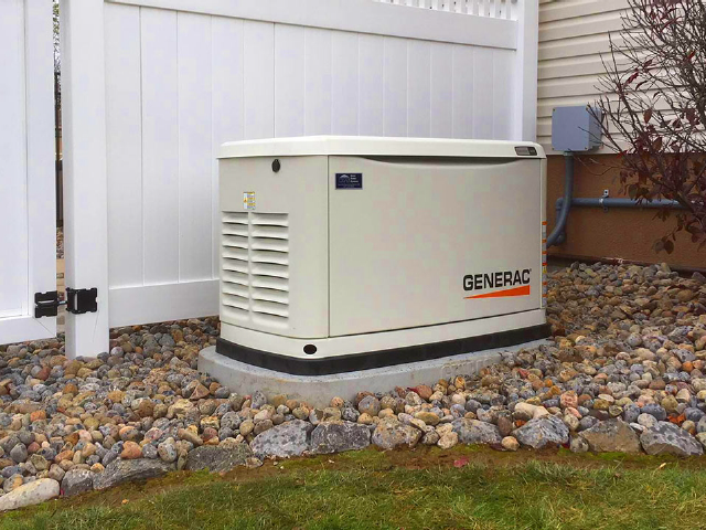 A generator is sitting in the grass next to a white fence.