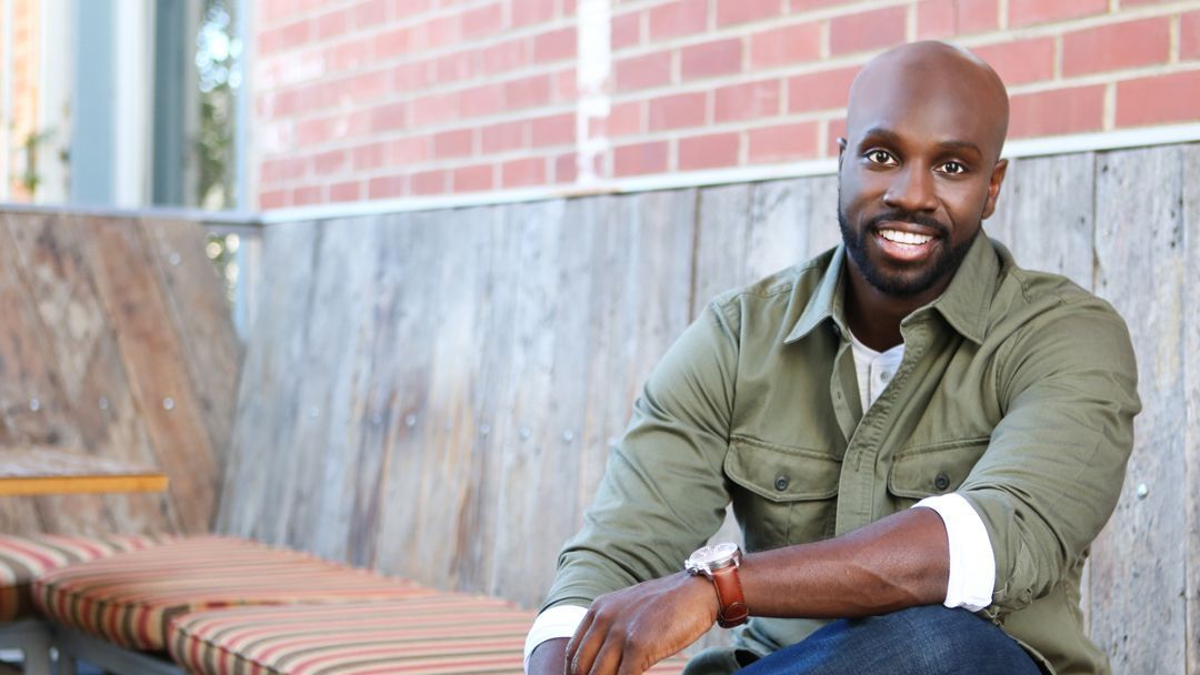 A bald Juntae Delane is sitting on a wooden bench in front of a brick wall.