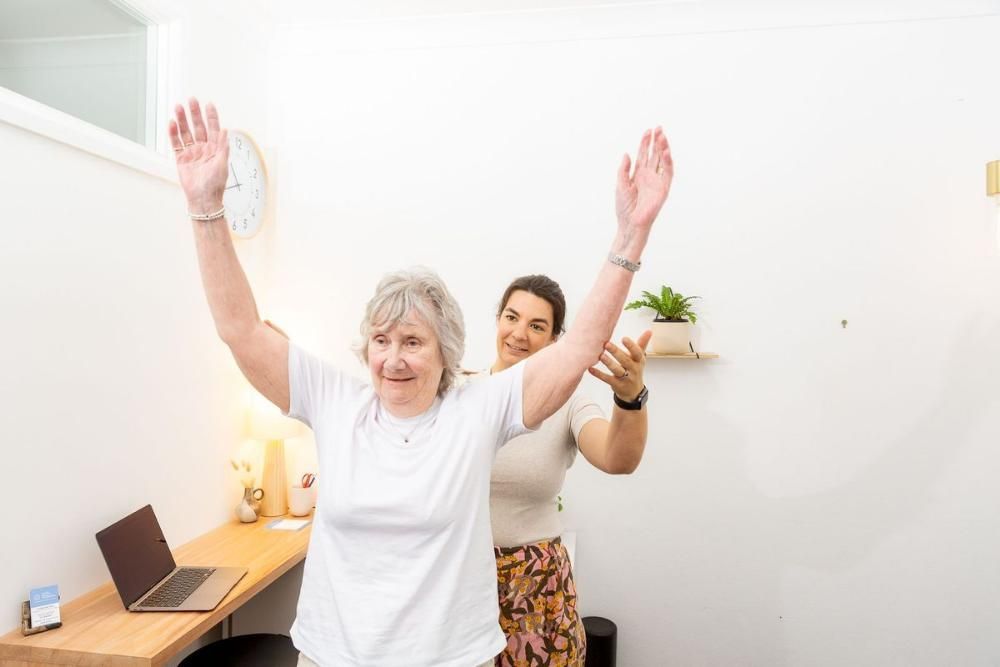 A Woman is Helping an Older Woman Stretch Her Arms in a Room — Align Health Therapies in Woonona, NSW