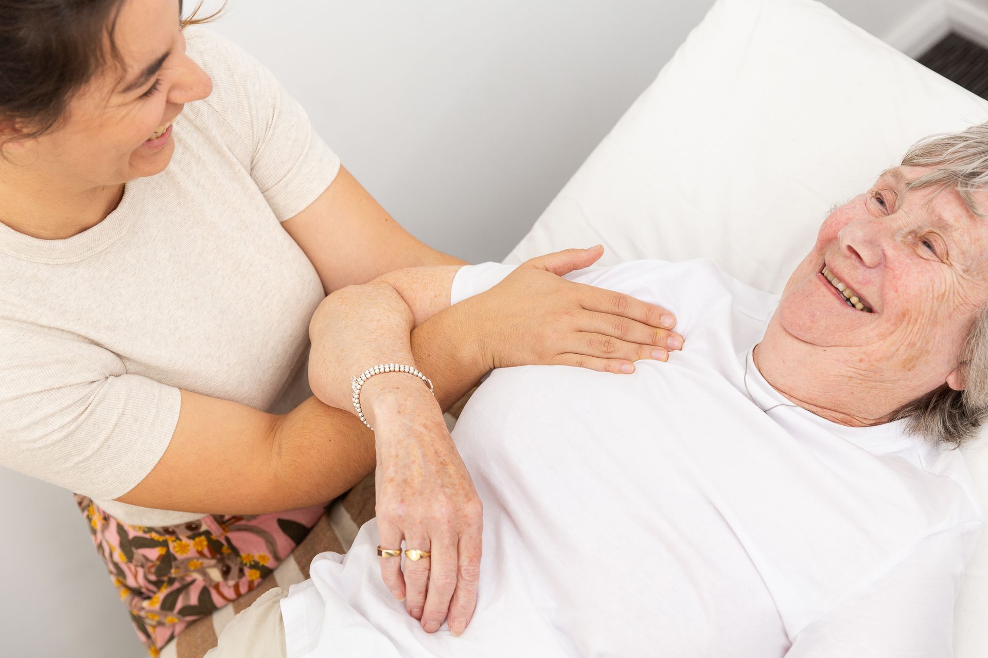 A Woman is Holding the Arm of an Elderly Woman Laying in a Bed — Align Health Therapies in Woonona, NSW