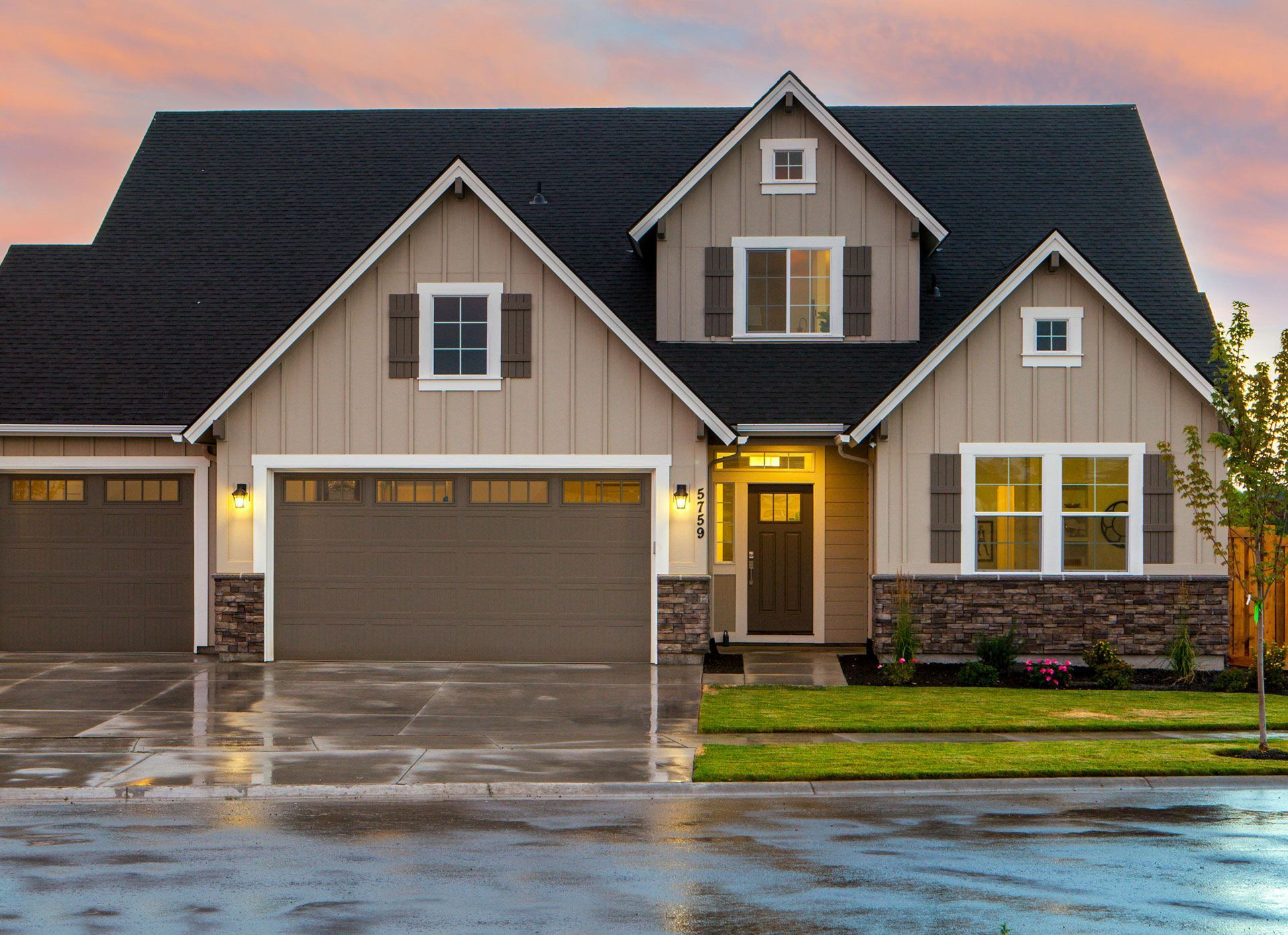 Two-story beige house with dark roof, brown garage doors, and rain-slicked driveway under a colorful sunset.