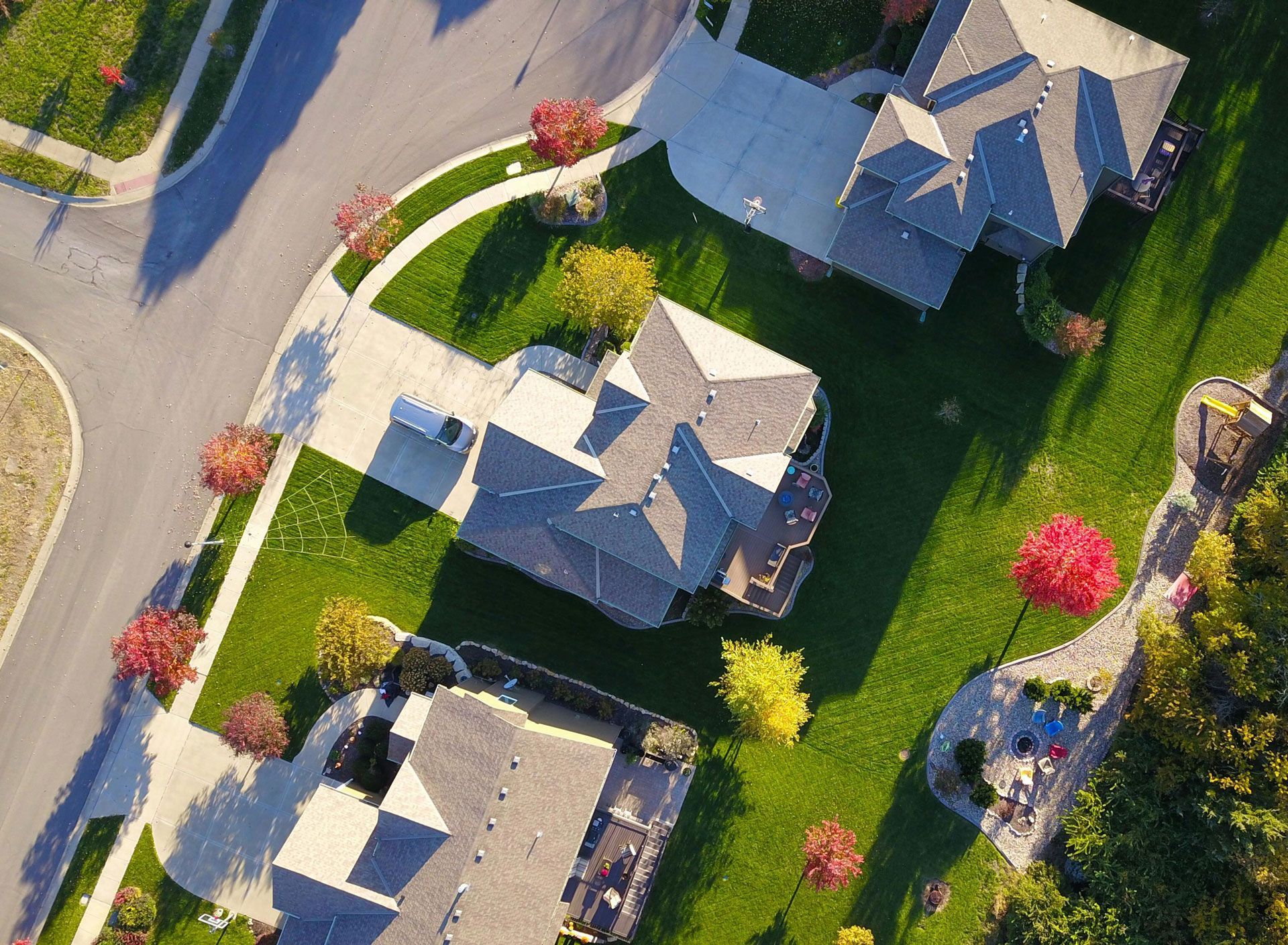 Aerial view of suburban homes with green lawns, trees, and driveways. Sunlight casts shadows.