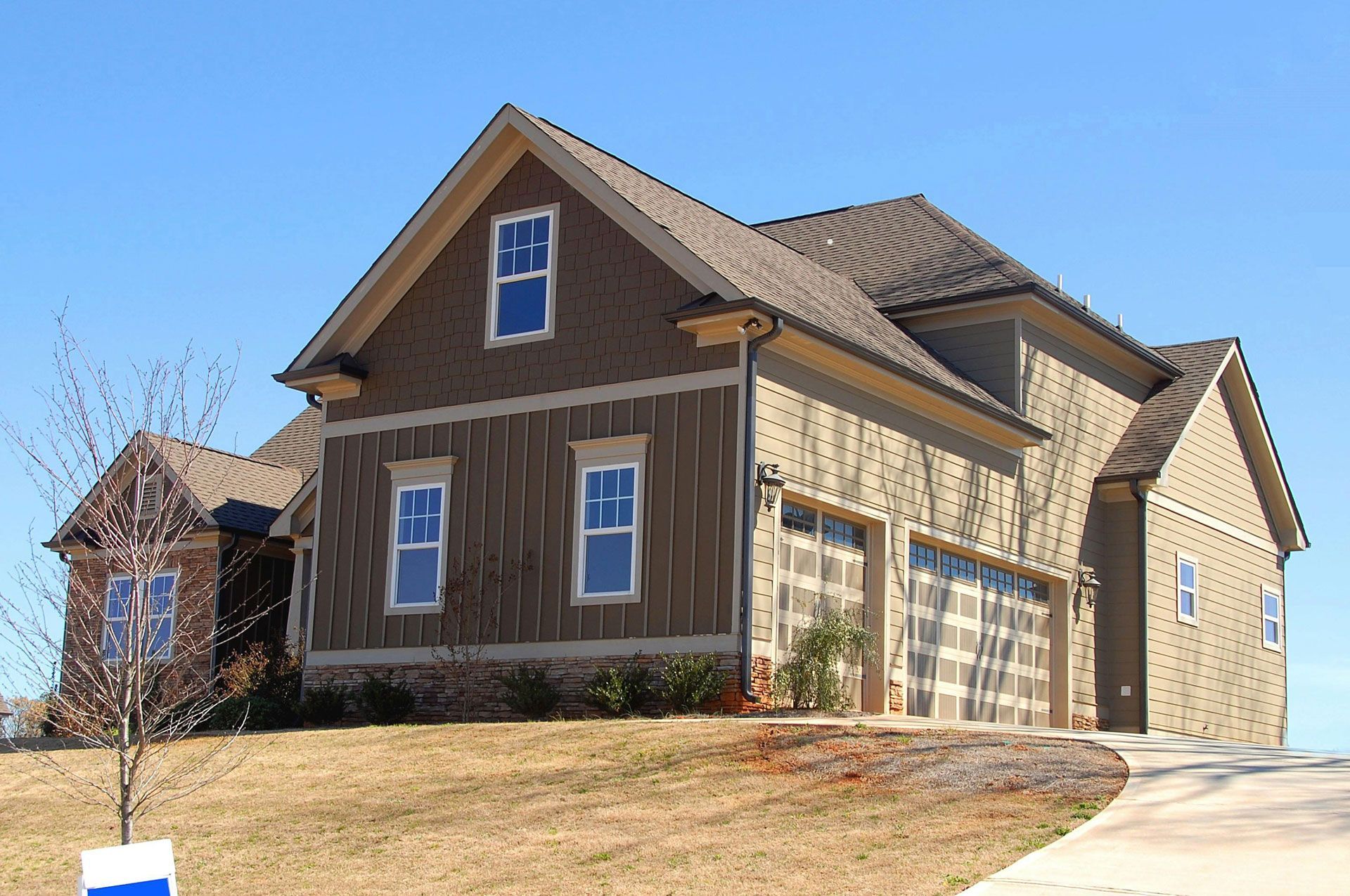 A brown and tan house with a garage and a driveway on a hill under a clear blue sky.
