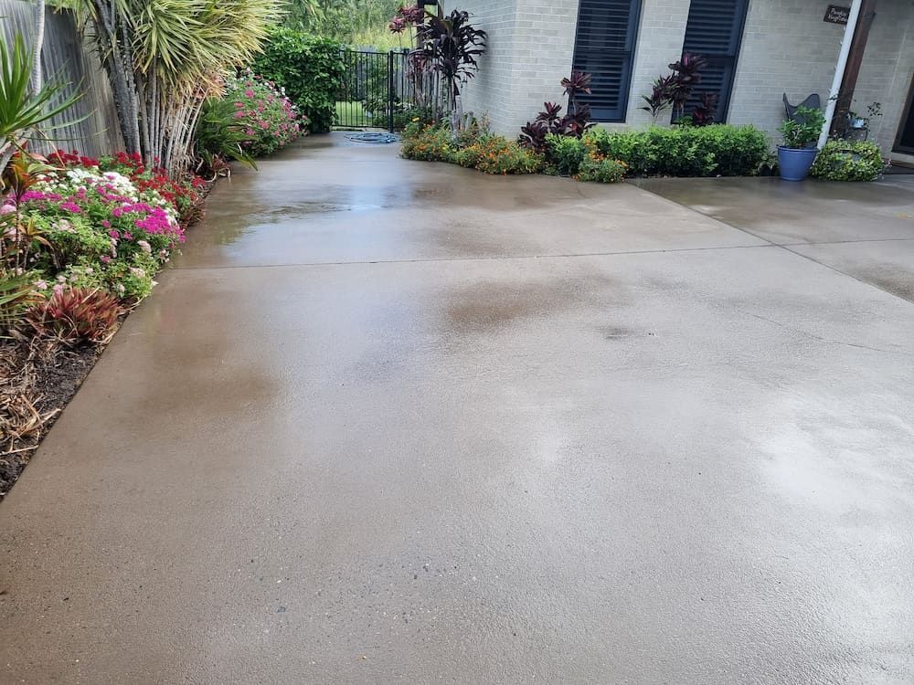 A Concrete Driveway Leading To A House With Flowers In The Background — Cap Coast Pressure Cleaning in Lammermoor, QLD