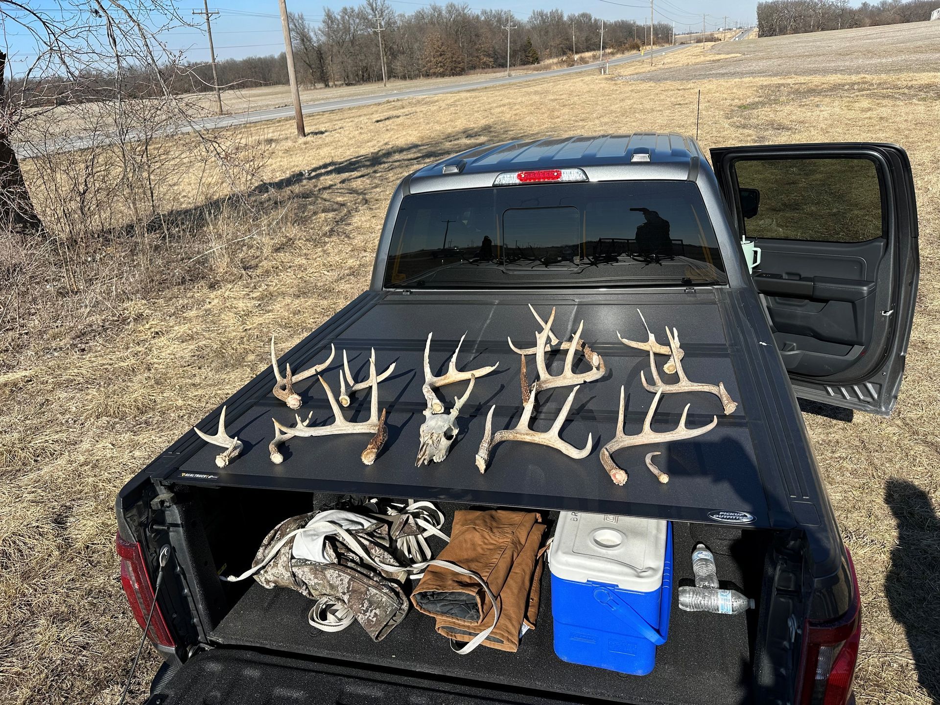 Pickup truck bed displaying multiple deer antlers, gear, and cooler in a rural setting.