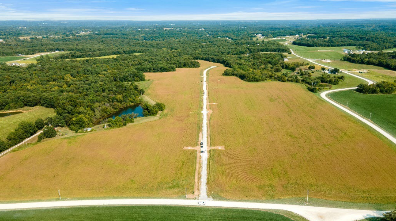 Aerial view of a field with a road, surrounded by trees and other fields under a blue sky.
