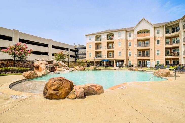 Swimming pool with lounge chairs in front of a multi-story apartment building, under a clear blue sky.