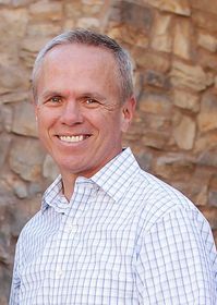 A man in a plaid shirt is smiling in front of a stone wall.