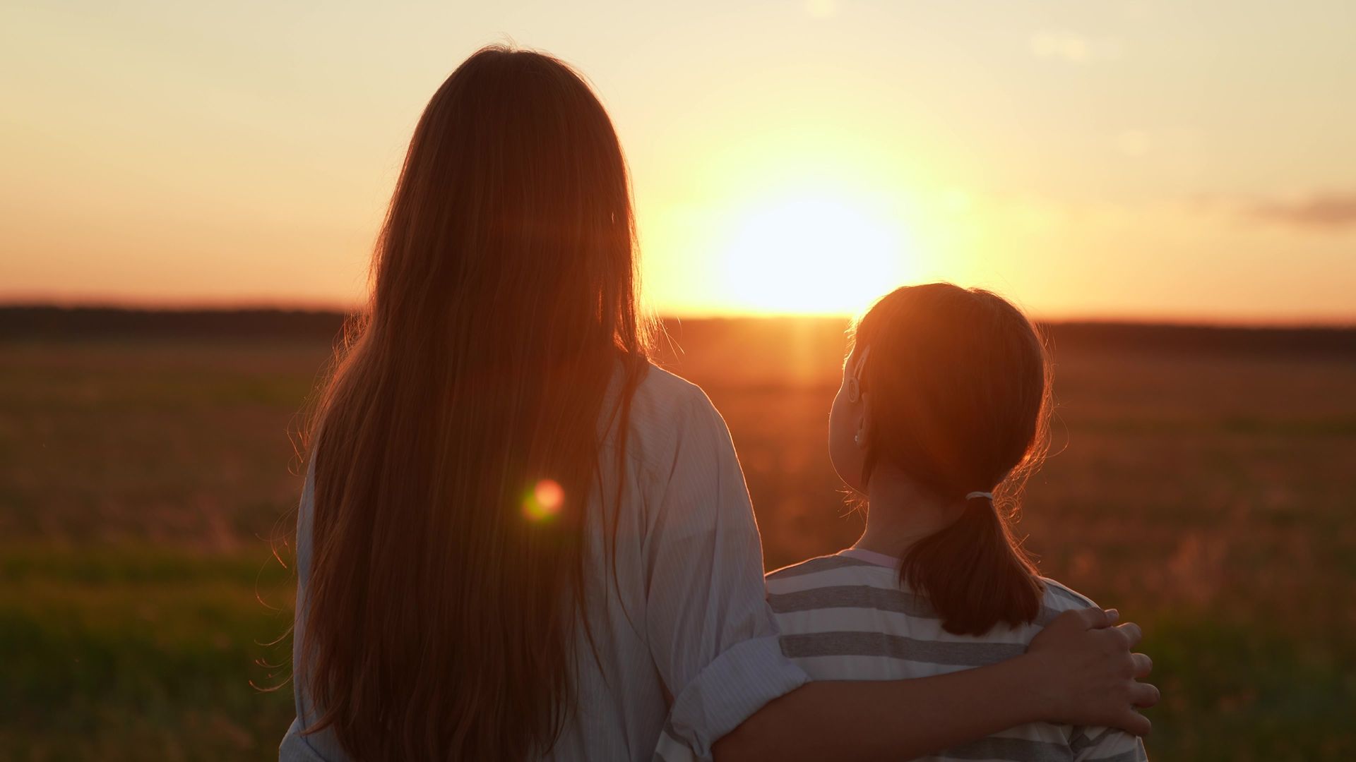 Two people watching a sunset over a field. One has an arm around the other's shoulder.