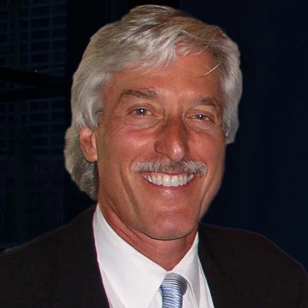 A man with gray hair and a mustache is wearing a suit and tie and smiling.