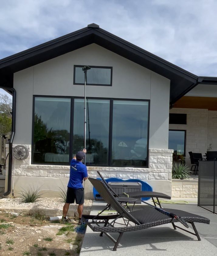 Man washing windows with a long-handled tool on a house with dark trim and a swimming pool in the background.