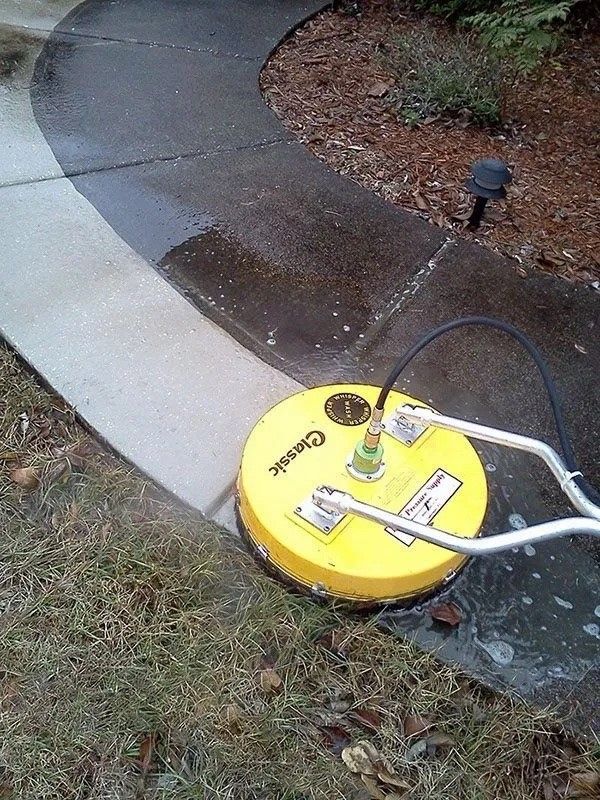 Yellow circular surface cleaner on a concrete walkway, cleaning a dark area to a light gray.