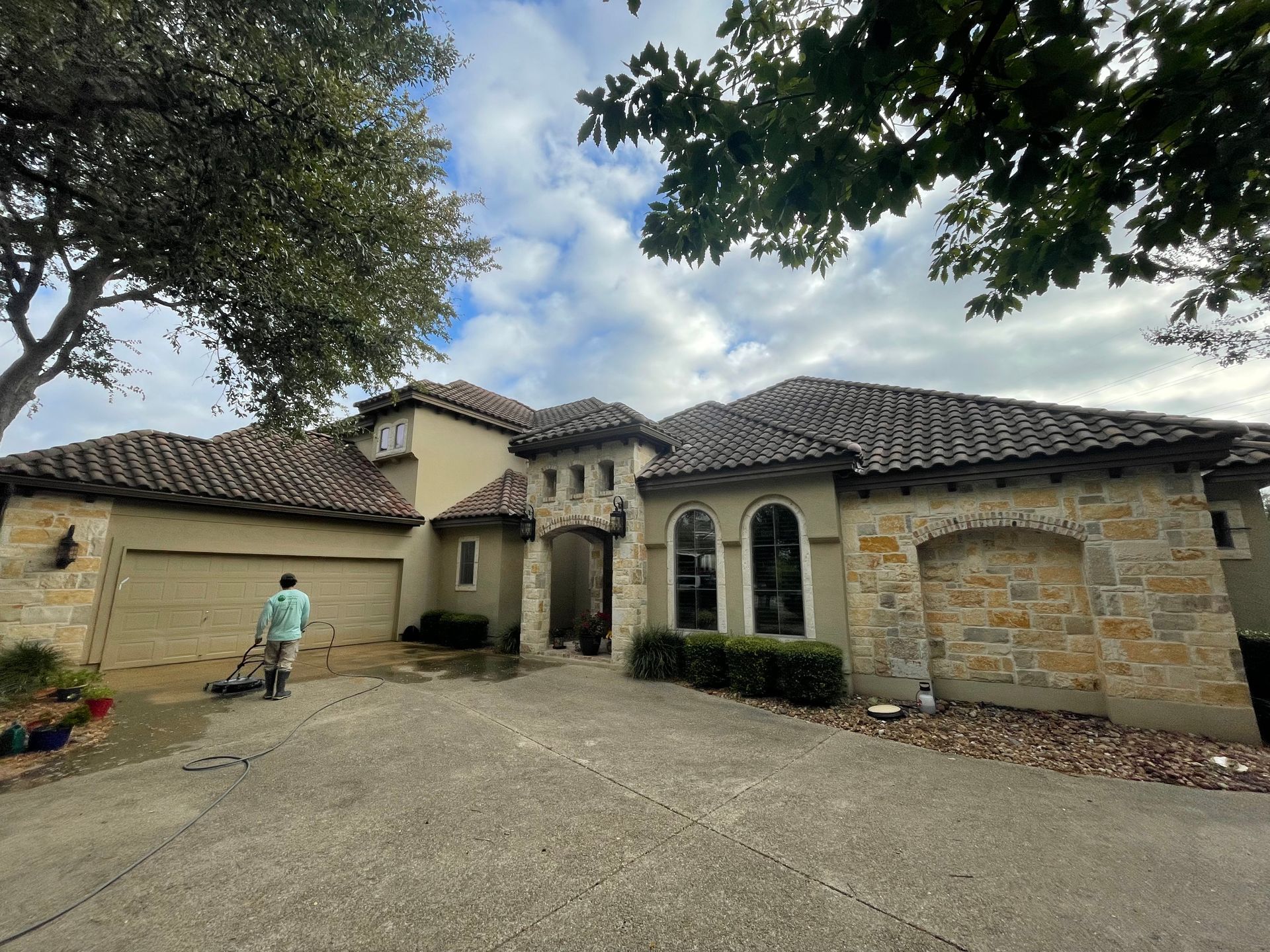 Man power washes the concrete driveway of a beige stone and stucco house with a tile roof under a cloudy sky.