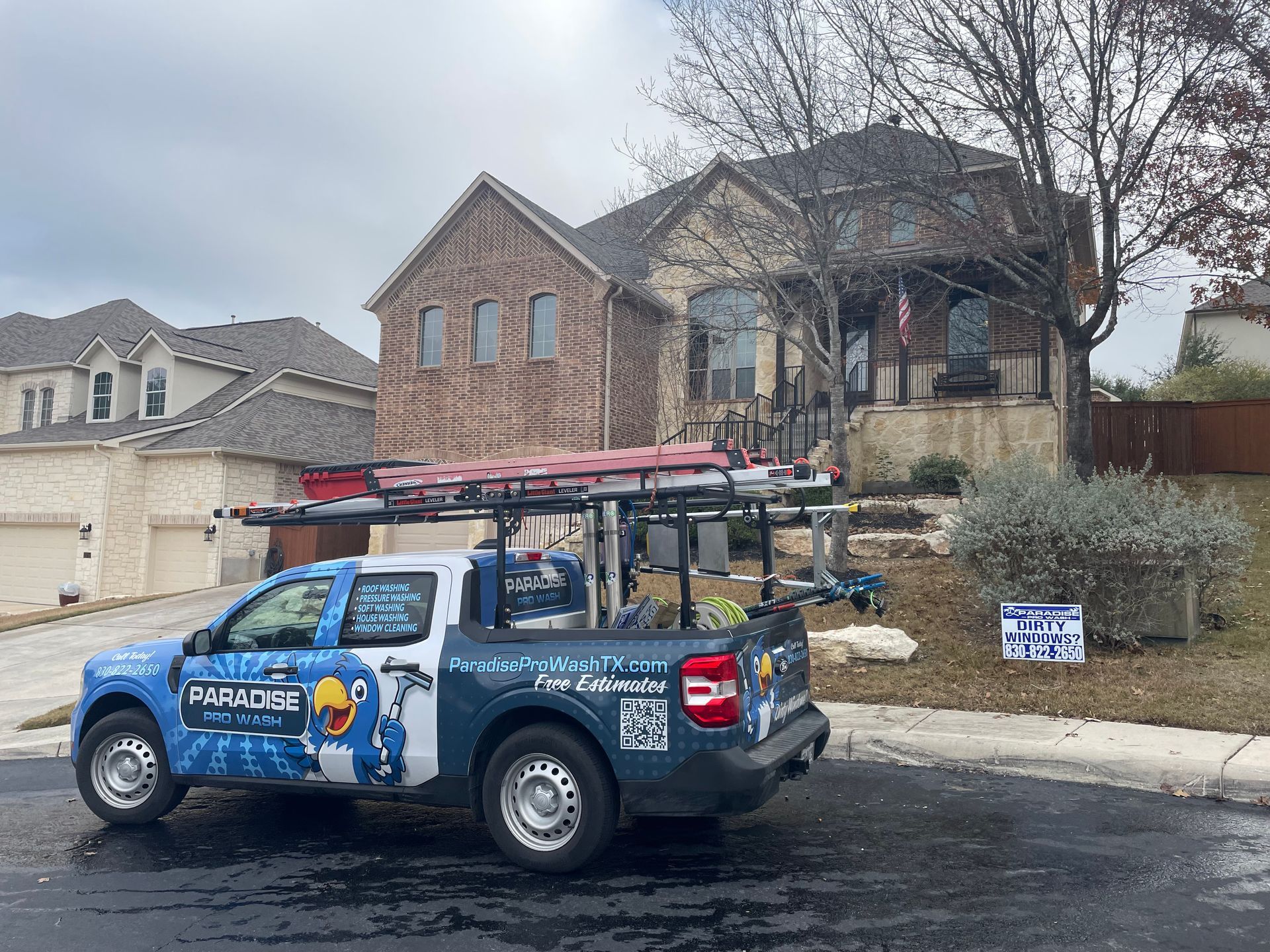 A blue and white HVAC service truck is parked on a suburban street in front of a brick two-story house.
