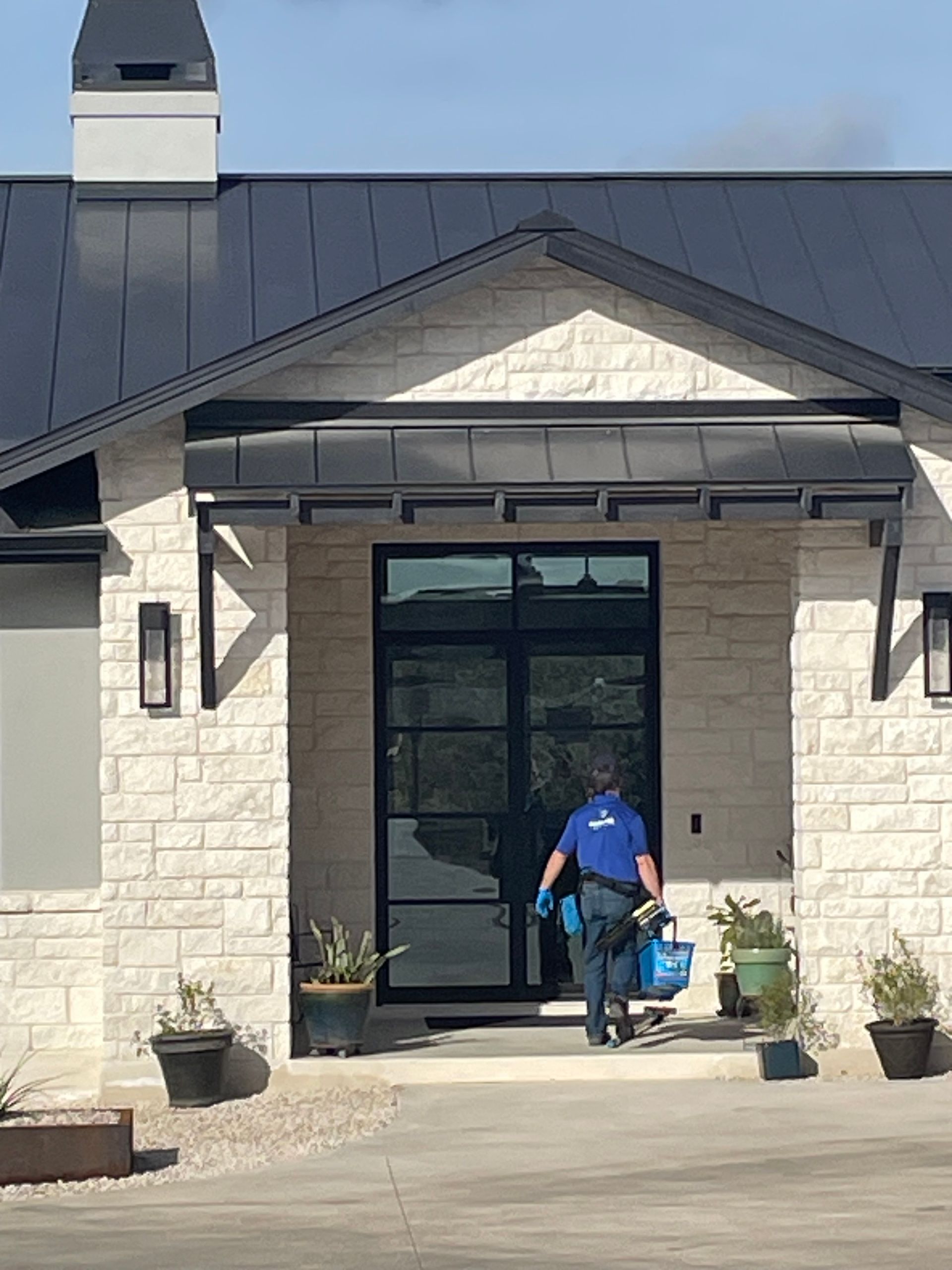 Person in blue shirt carrying cleaning supplies enters a house with stone facade, dark roof, and door.