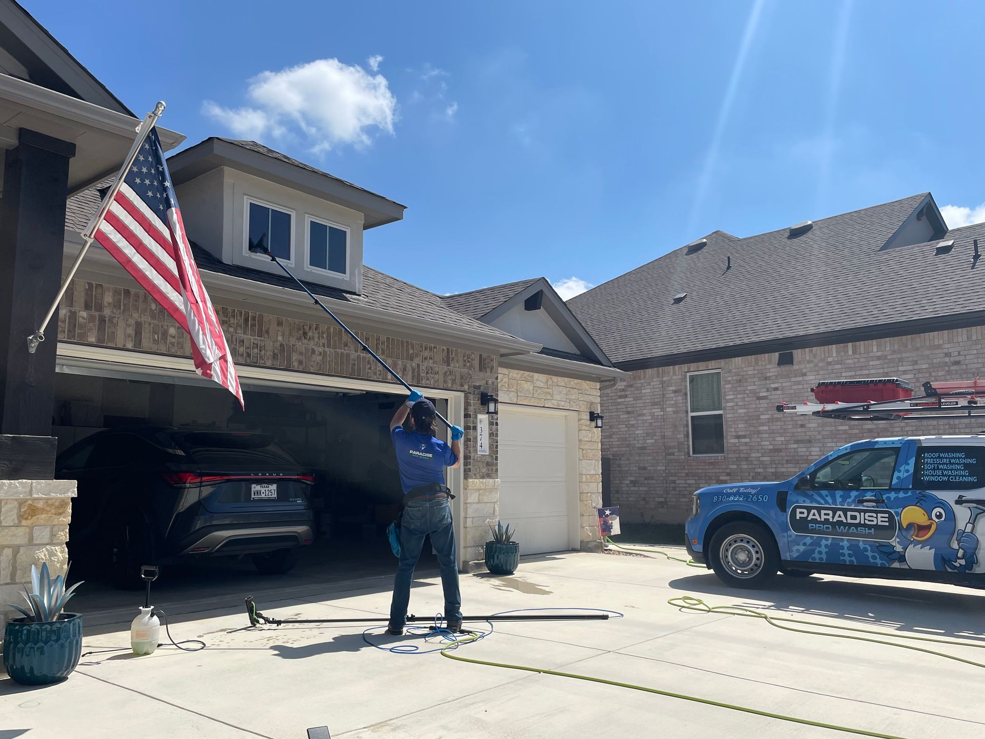 A worker uses a long-reach pole to clean an upper-story window on a brick house with a parked car and a service truck.