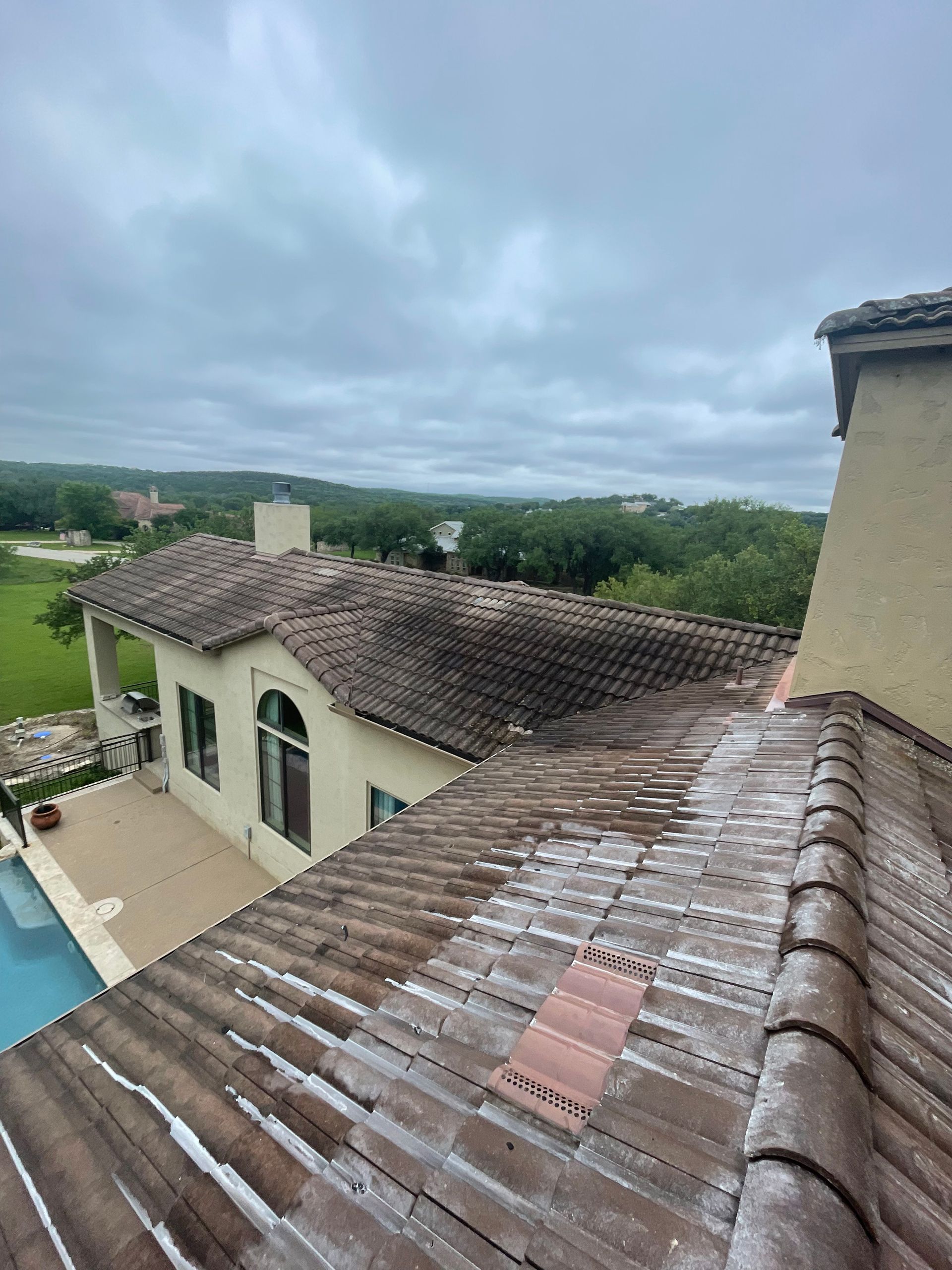Rooftop view of a villa with terracotta tiles, pool, and cloudy sky over green trees