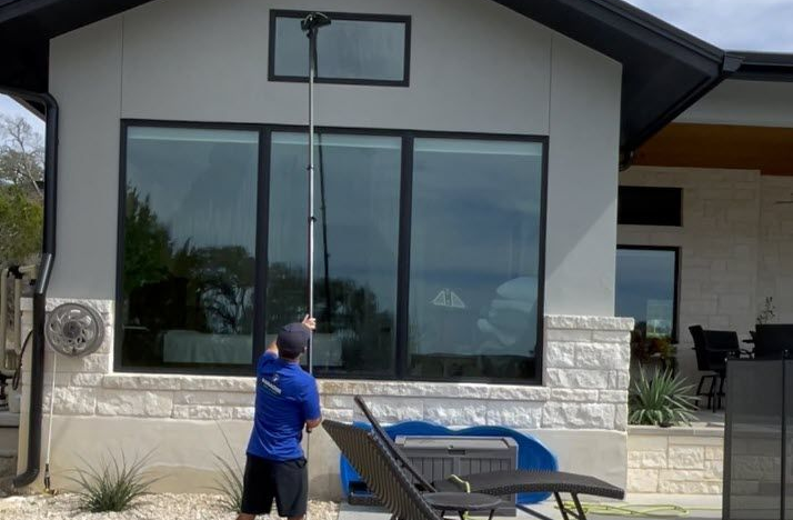 Person cleans large window with a telescoping pole. Building has stone and white siding in Bexar County, TX.