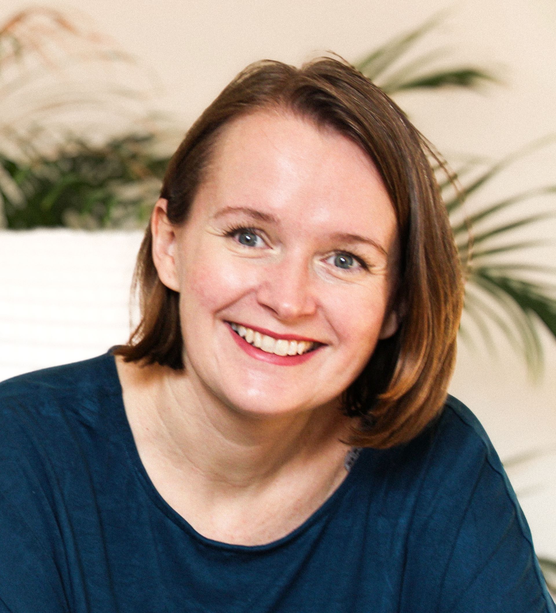 Eine Person mit kinnlangem braunem Haar und einem freundlichen Lächeln, die ein dunkelblaues Oberteil trägt, vor einem weichen, blattreichen Hintergrund.