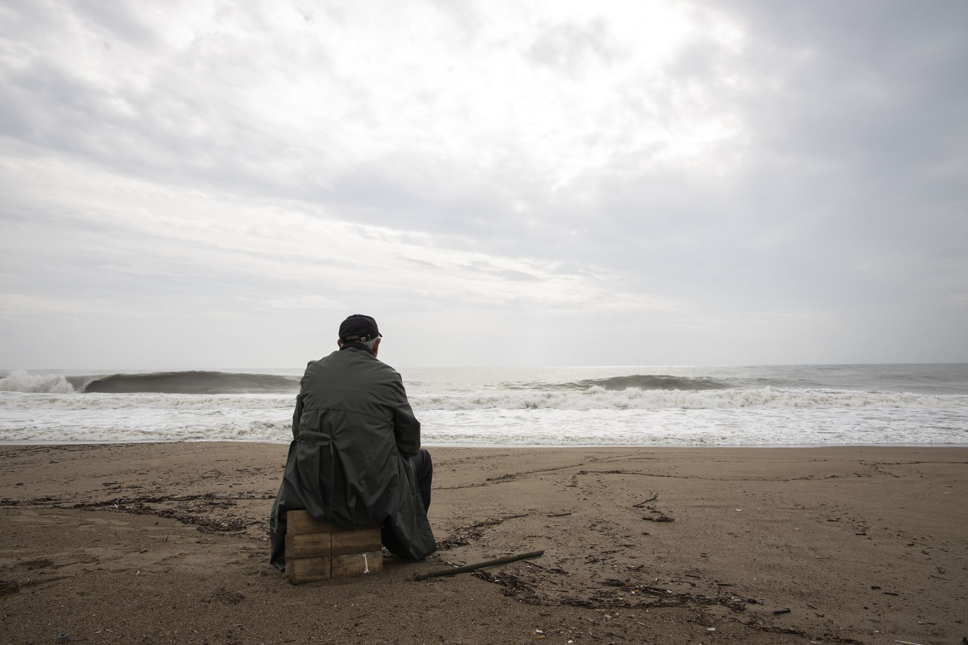 Eine Person in dunklem Mantel und Hut sitzt auf einer Kiste an einem Sandstrand und blickt unter einem bewölkten Himmel aufs Meer hinaus.