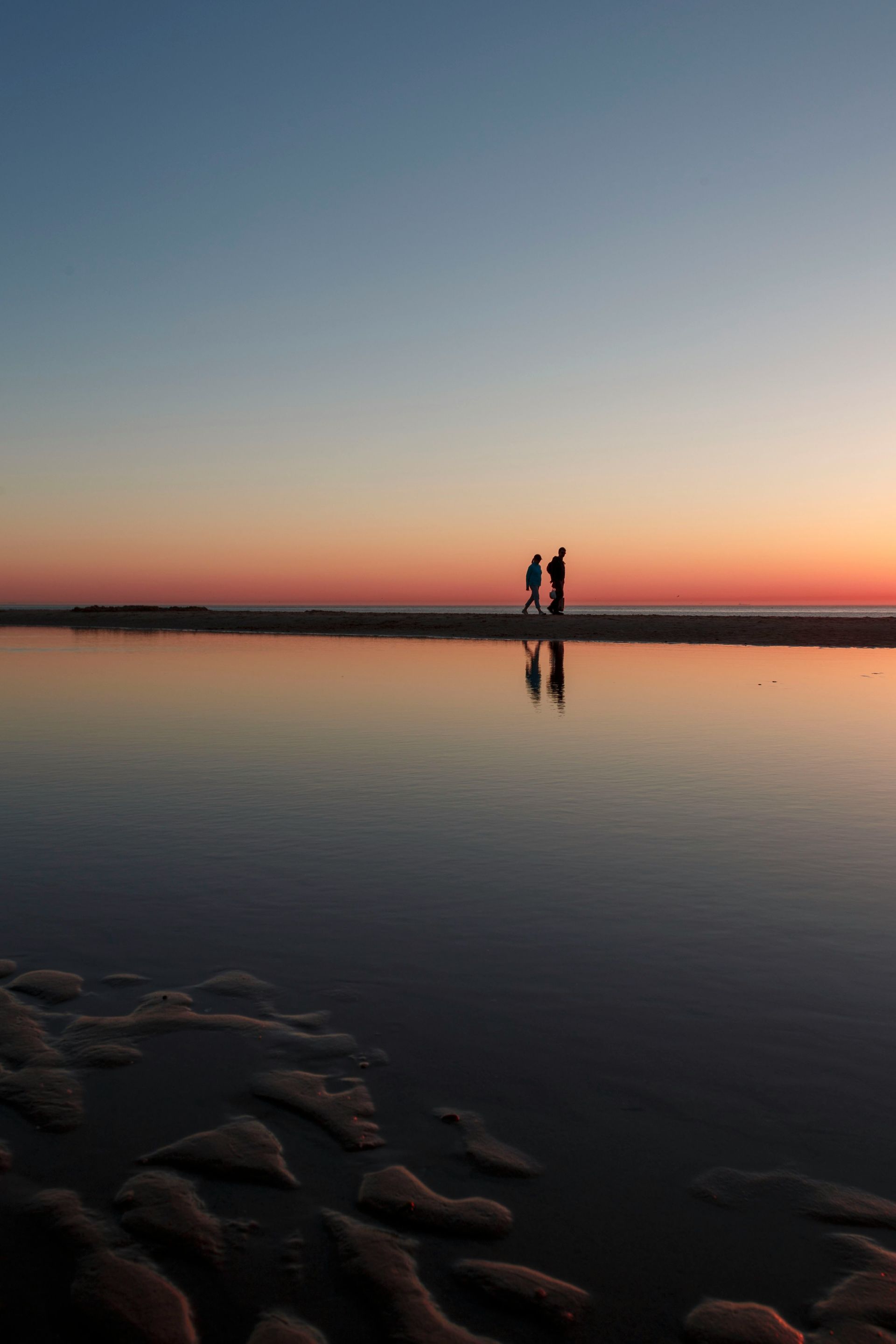 Zwei Gestalten stehen bei Sonnenuntergang auf einer Sandbank, die sich im ruhigen, flachen Wasser spiegeln, im Vordergrund ist der gekräuselte Sand zu sehen.