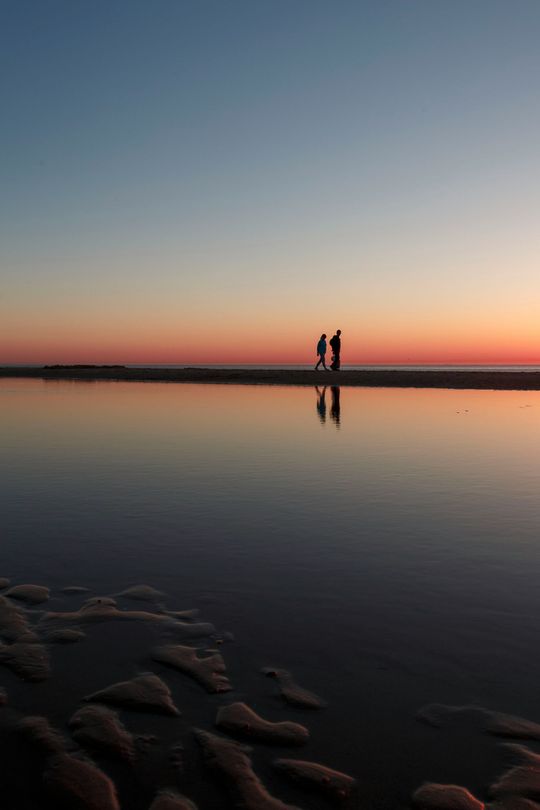 Zwei Gestalten stehen bei Sonnenuntergang auf einer Sandbank, die sich im ruhigen, flachen Wasser spiegeln, im Vordergrund ist der gekräuselte Sand zu sehen.