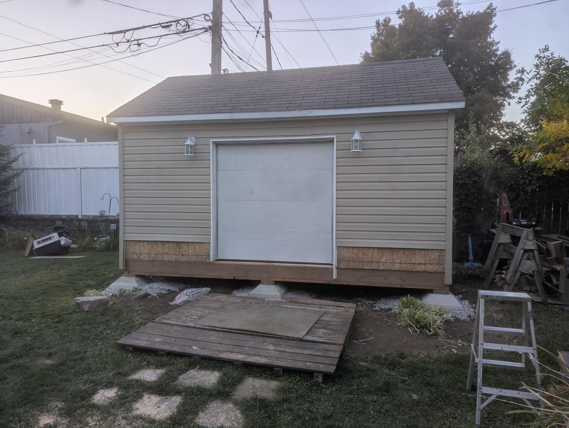 Abri beige avec porte blanche, deux lampes, toit gris, sur une cour herbeuse, rampe en bois et échelle.