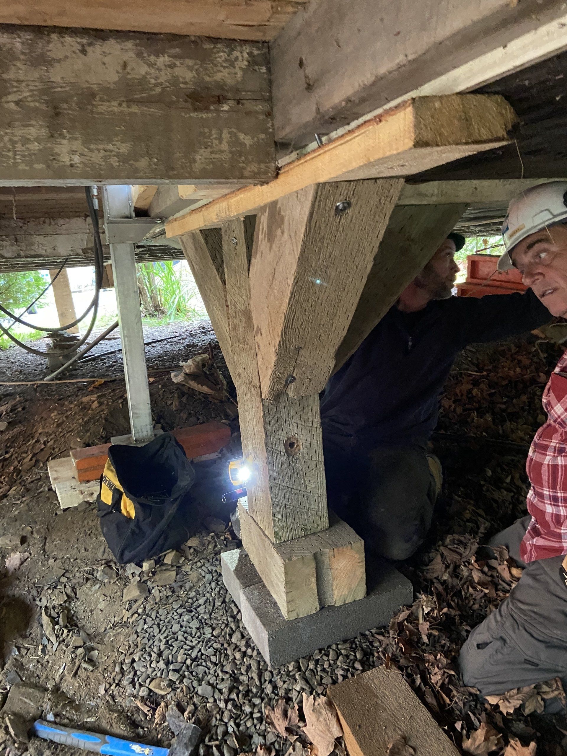 Un homme est agenouillé sous une maison et regarde les fondations.