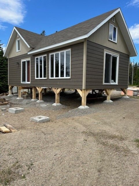 Une maison sur pilotis de style chalet, avec un bardage brun et des fenêtres à cadre blanc, située sur un terrain de gravier sous un ciel bleu.