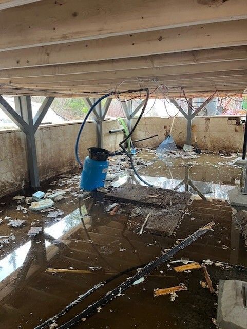 Sous-sol inondé sous une terrasse en bois, avec de l'eau, des débris et un conteneur bleu.