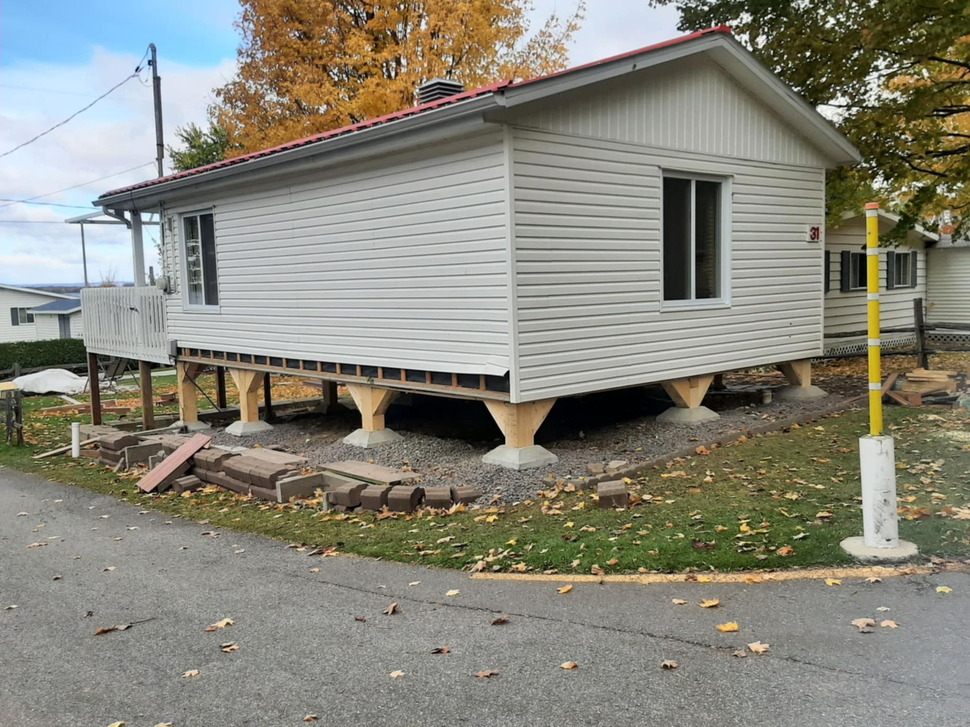 Cabane blanche sur pilotis, posée sur du gravier, avec un toit rouge. Feuilles d'automne sur les arbres en arrière-plan.