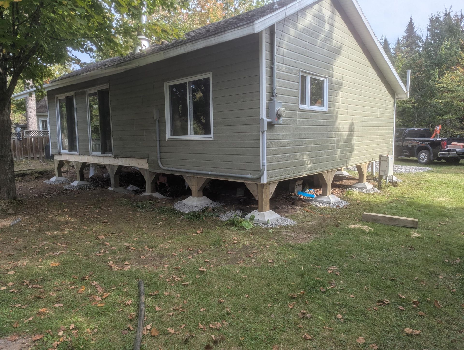 Maison à façade verte sur pilotis en bois surélevés, avec deux fenêtres et des portes coulissantes en verre, située dans une cour gazonnée.