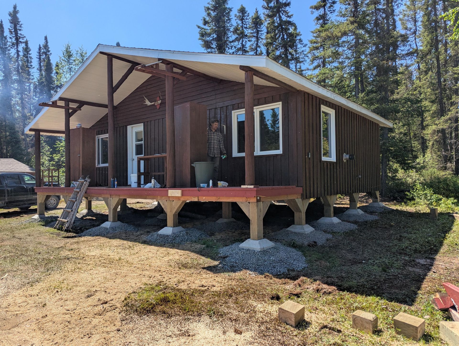 Cabane brune sur pilotis avec porche, entourée de gravier et d'arbres.