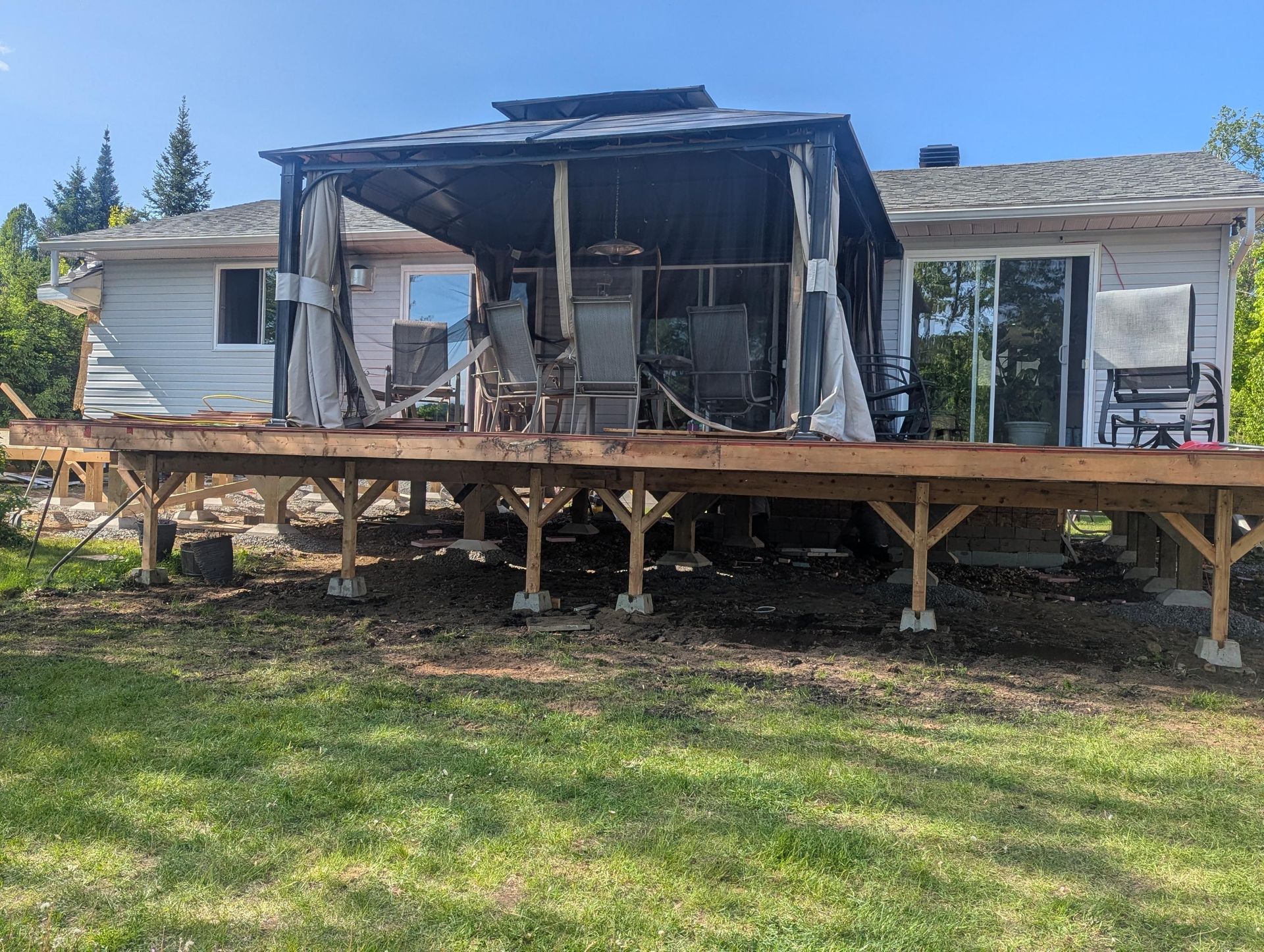 Terrasse en bois en construction avec un kiosque, attenante à une maison de couleur claire. Pelouse verte au premier plan.