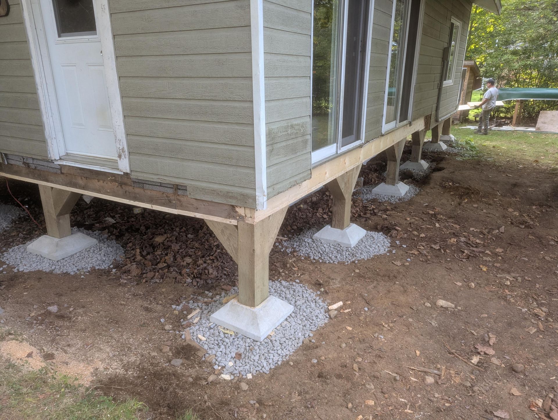 Bâtiment surélevé avec supports en bois sur dalles de béton, entouré de gravier et de terre.