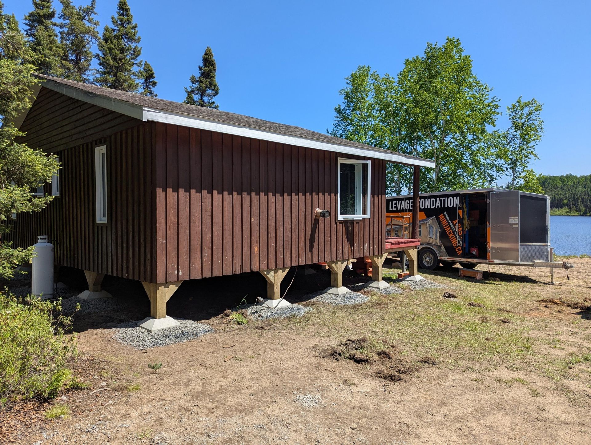 Cabane brune sur blocs de béton près de l'eau, avec une caravane garée à côté. Belle journée ensoleillée.